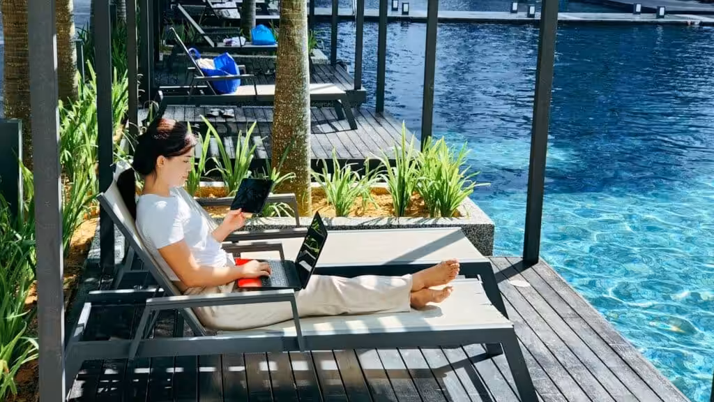 A woman working on a laptop and holding a tablet while relaxing on a lounge chair by a swimming pool, illustrating the digital nomad lifestyle.